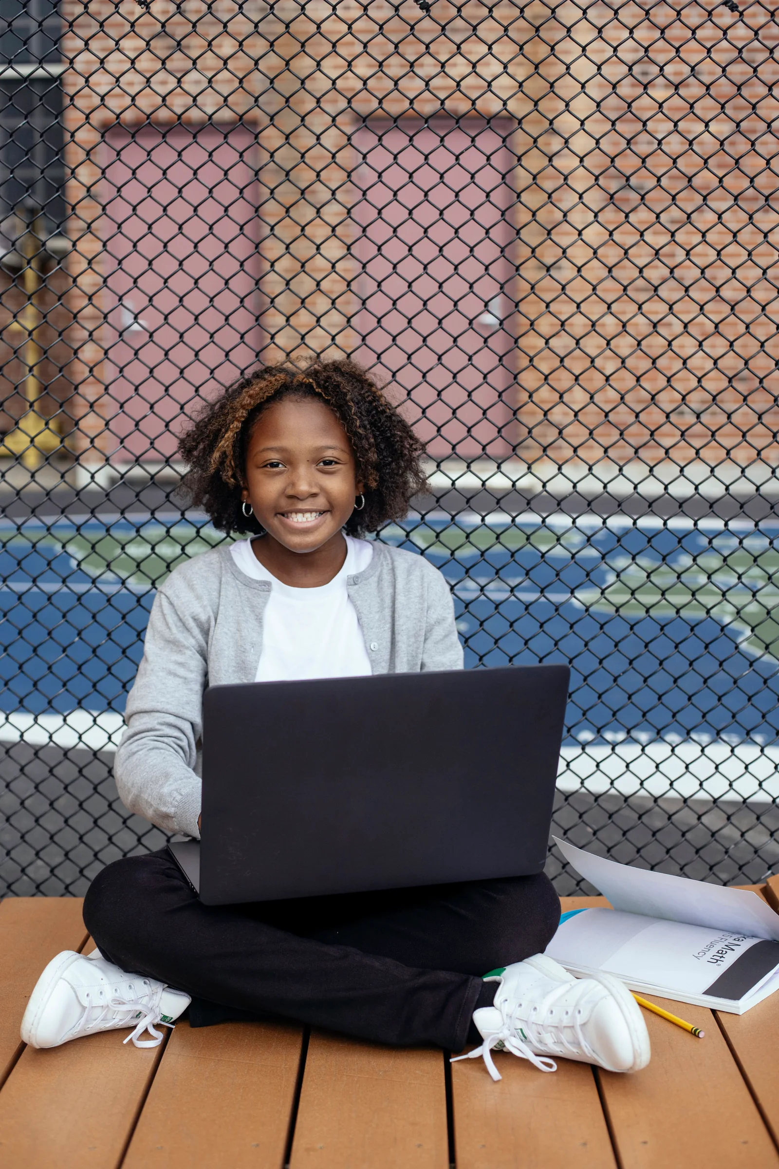 A smiling Black student seated with a laptop beside a schoolyard fence